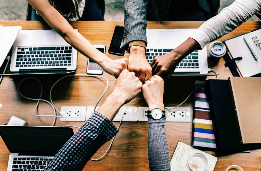 articles-632 - Cambridge Car Audio Topdown View of Diverse Hands in a Fistbump Circles over a Cluttered Desk with Laptops Phones and Notebooks Symbolizing Teamwork